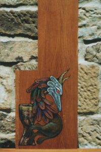 Pintura sobre madera en estilo medieval de un dragón con forma de S de perfil con cuernos, sin alas ni brazos, torso emplumado y patas de león protegiendo el grial