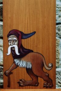 Pintura sobre madera en estilo medieval de un anciano con gorro morado, largos bigotes blancos, cuerpo de león y un solo brazo apoyado en un batón con forma de tau