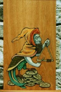 Pintura sobre madera en estilo medieval de un mercader judío de perfil con un gabán con capucha color amarillo y naranja, cuerpo de pájaro en tonos verdosos, erguido sobre una montaña de monedas de oro con un pentagrama cincelado y tocando un triángulo dorado