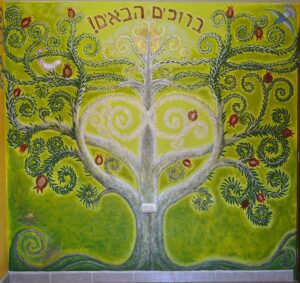 Mural artístico del Árbol de la Vida con elementos simbólicos como granadas, aves y espirales, acompañado del texto hebreo “Bienvenidos”.