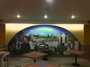 Mural de gran formato que representa una plaza imaginaria de la colonia San Ángel en Ciudad de México, ambientado en unos 1930 alternativos, con escenas cotidianas y figuras en proceso de creación. Obra que fusiona arte urbano, narrativa visual y cultura local en un entorno arquitectónico